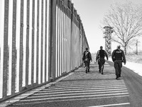Black and white BW image of border police walking alongside the fortification fence with a watch tower post in the background for police and...