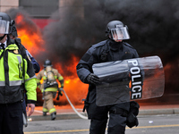Protected by police officers in riot gear, firefighters battle a car fire, set to a limousine after protests erupt after Donald Trump become...