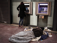 A homeless man lies on the pavement in front of a bank while a woman withdraws money from the ATM in Athens on January 18, 2015. (