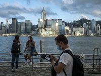Two Women Chat and a man looks at his phone in front of the Hong Kong City Skyline in Hong Kong, Monday, May 10, 2021. (