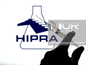 In this photo illustration, a close up of a hand holding a medical syringe in front of the Hipra, S.A. logo (