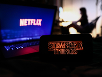 Stranger Things series logo displayed on a phone screen, Netflix logo displayed on a laptop screen and a silhouette of a woman in the backgr...