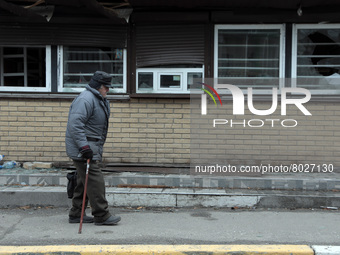 KYIV REGION, UKRAINE - APRIL 06, 2022 - An elderly man with a cane passes by a building with shattered windows as a result of shelling by th...