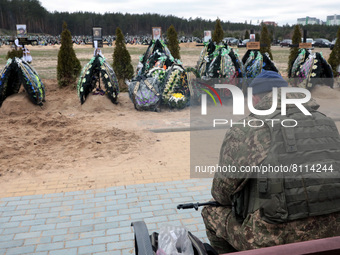 IRPIN, UKRAINE - APRIL 20, 2022 - An armed soldier in a camouflage uniform and bulletproof vest faces the graves of those who perished in th...