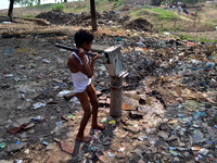 An indian boy pulls water in hand pump in a A highly polluted compound with plastic bags and other garbage is next to a shanty town, in Alla...