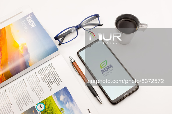 In this photo illustration an Archer Daniels Midland (ADM) logo seen displayed on a smartphone screen on a desk next to a cafe, a pen, glass...