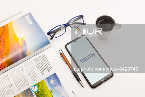 In this photo illustration a Truth Social logo seen displayed on a smartphone screen on a desk next to a cafe, a pen, glasses and a magazine...