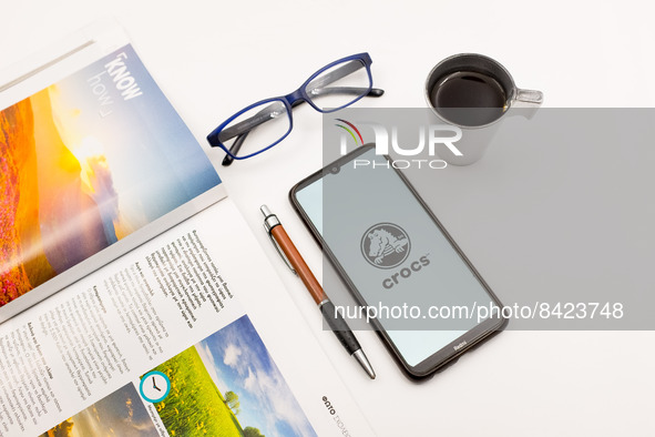 In this photo illustration a Crocs logo seen displayed on a smartphone screen on a desk next to a cafe, a pen, glasses and a magazine in Ath...