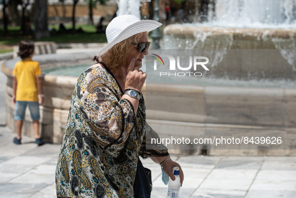 A woman wearing a hat for protection and holding a bottle of water walks past a fountain at Syntagma Square during a heatwave in Athens, Gre...