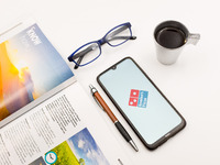 In this photo illustration a Domino’s Pizza logo seen displayed on a smartphone screen on a desk next to a cafe, a pen, glasses and a magazi...