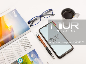 In this photo illustration a Reebok logo seen displayed on a smartphone screen on a desk next to a cafe, a pen, glasses and a magazine in At...