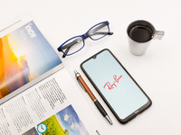 In this photo illustration a Ray-Ban logo seen displayed on a smartphone screen on a desk next to a cafe, a pen, glasses and a magazine in A...