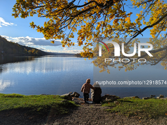 Mother And Child Sit By Lake Shoreline In Autumn Sunlight