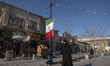 A veiled Iranian woman is walking past an Iranian flag that is hanging on a sidewalk in th...