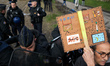 A protester is being pushed away by riot police while holding a placard against the A69 hi...