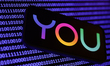 A binary code displayed on a laptop screen and You logo displayed on a phone screen are se...