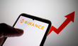 The Binance logo appears on a smartphone screen, and an exchange rate arrow appears on a c...