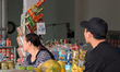 A woman and a man sit at a sidewalk shop selling packaged snacks, bananas, and papayas in...
