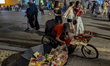 A street vendor arranges colorful handmade pop-up cards on a tricycle cart along a busy pe...