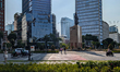 People cross a street near a statue of a historical figure in front of modern office build...