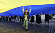 A boy in a traditional Ukrainian embroidered shirt stands under the flag of Ukraine on Der...