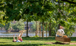 People spend a sunny afternoon by the Vltava River under the shade of green trees in Pragu...