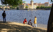 A group of people stands on the sandy bank of the Vltava River, while a man stands nearby...
