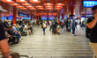 People sit and walk in the main corridor of Prague Central Station, surrounded by signage,...