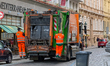 Garbage collectors operate a waste collection truck on a cobblestone street in the city ce...