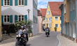 A group of motorcyclists rides through a colorful and narrow street lined with traditional...