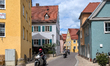 A group of motorcyclists rides through a colorful and narrow street lined with traditional...