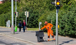 A street cleaner in a high-visibility orange uniform sweeps the pavement and pushes a tras...