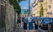 Three women wearing hijabs walk along a street lined with parked cars and shops in Berlin,...