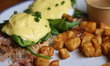 Eggs Benedict with ham and maple syrup glazed bacon is served on a bun with a side of pota...