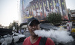 An Iranian man smokes an e-cigarette while standing beneath a giant banner featuring symbo...