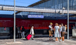 Passengers stand with luggage, and a group in traditional Bavarian clothing gathers on the...