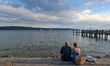 A man wearing a hat and a woman in a blue dress sit together at the waterfront with ducks...