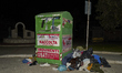 A full recycling bin for used clothes overflows with donations and is surrounded by garbag...