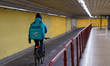 A food courier wears a bright blue jacket and a large Deliveroo backpack and rides a bicyc...