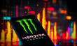The Monster Energy icon is displayed on a mobile phone with a financial stock graph seen i...