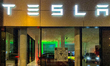 An illuminated facade of a TESLA sales showroom features large windows displaying current...