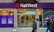 Pedestrians walk past the illuminated storefront of a NatWest bank branch located at 16 No...