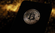 A Bitcoin coin appears on a smartphone screen with a black and gold illustration in the ba...