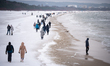 People walk along a snow-covered beach with Sopot visible in the distance along the coastl...
