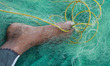 A fisherman uses his leg to hold a fishing net as he repairs it under a tree on a hot summ...