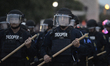 Dozens of St. Louis riot police armed with batons march toward a large group of protesters...