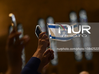An attendee is holding up an Israeli flag lit by a phone while a standing-room crowd gathers as Governor Josh Shapiro and Senator Bob Casey...
