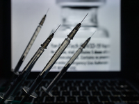 An illustrative image of medical syringes seen in front of an image of a vial containing Pfizer-BionTech vaccine displayed on a screen.
On M...