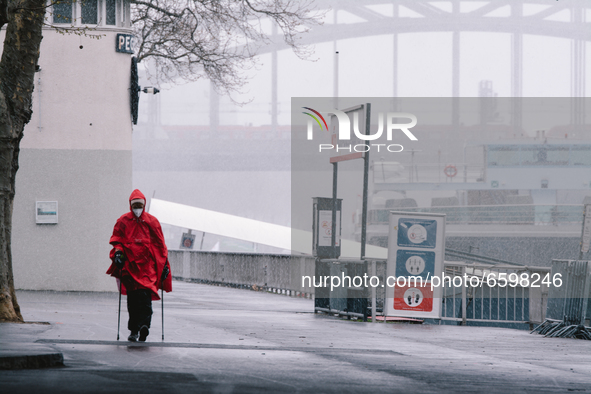 a woman with raincoat walking along the rhine river as rare April snowfalls in Cologne, Germany on April 6, 2021 