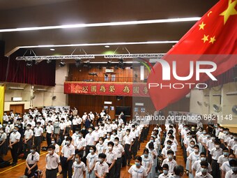 A Flag of China on a Flag Pole next to Students wearing face masks standing under a banner with patriotic slogans, during a flag raising cer...